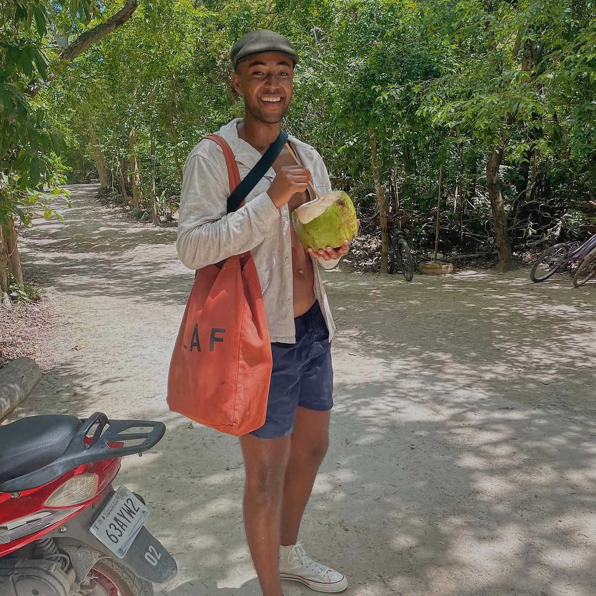 Person holding a coconut in a forested area with an orange bag and a motorcycle in the background.