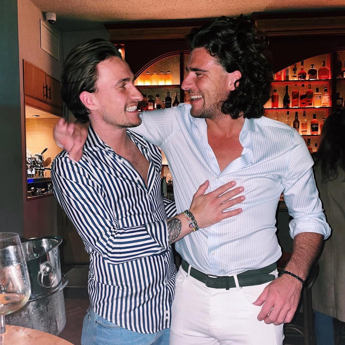 Two men embracing in a bar setting with shelves of bottles in the background.